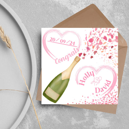 square wedding card with a bottle of champagne bursting with hearts in varying shades of pink. Above the bottle is a heart that holds the date and says contrats, and below is a repeated heart with the names of the couple inside