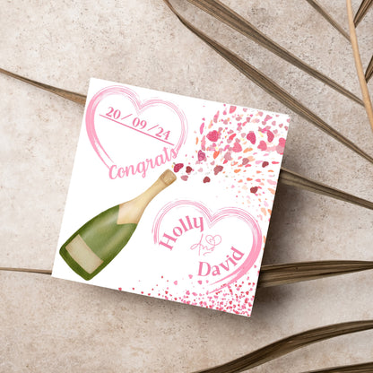 square wedding card with a bottle of champagne bursting with hearts in varying shades of pink. Above the bottle is a heart that holds the date and says contrats, and below is a repeated heart with the names of the couple inside