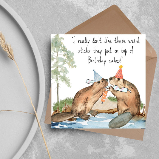 Image of a square birthday card with a watercolour image of 2 beavers with party hats on, chewing candles and handwritten style text  above them that reads - i really don&#39;t like these weird sticks they put on top of birthday cakes!