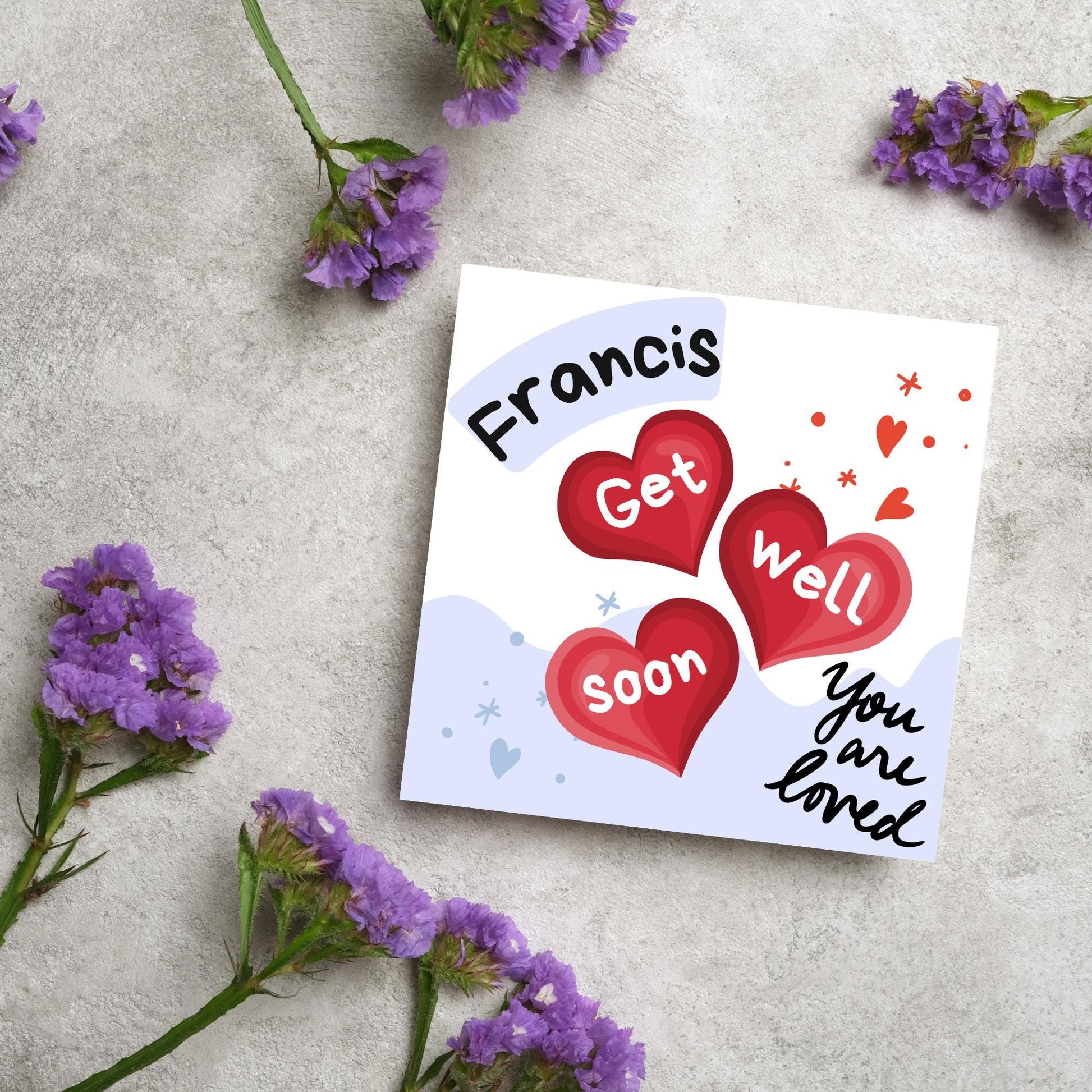 square card with white and blue background. 3 red hearts are in the centre of the card with get well soon overlayed in white, the top corner has curved text with the recipients name and in the bottom corner the text you are loved in black cursive
