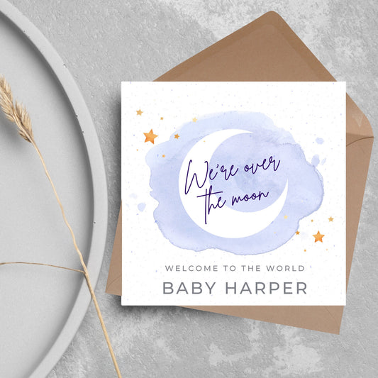 square card with a watercolour light blue background with a white crescent moon and yellow stars with navy blue hand script that reads - we&#39;re over the moon, and at the bottom in grey text welcome to the world baby harper