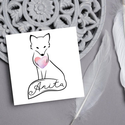 Square card with simple black line drawing of a fox  sitting with its tail wrapped around its feet. It has a pink watercolour heart on its chest and personalised with a name on the tail in a calligraphy font