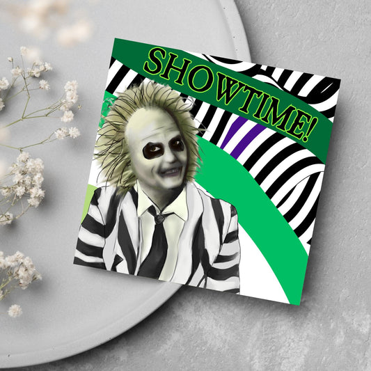 square greeting card with a drawn image of beetlejuice grinning in from of a black, green and purple striped background. On one of the biggest stripes is the word showtime in bold capital letters in a font similar to the movie title
