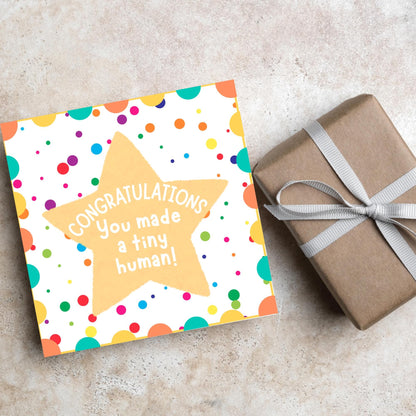 Image of a square card, the card design is a multicoloured polka dot design with a large yellow coloured star in the middle and white text against the star that reads CONGRATULATIONS You made a tiny human!
