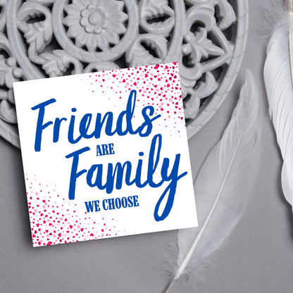 Square card with small hearts on opposite corners in shades of pink and blue writing in the centre that reads Friends are Family we choose