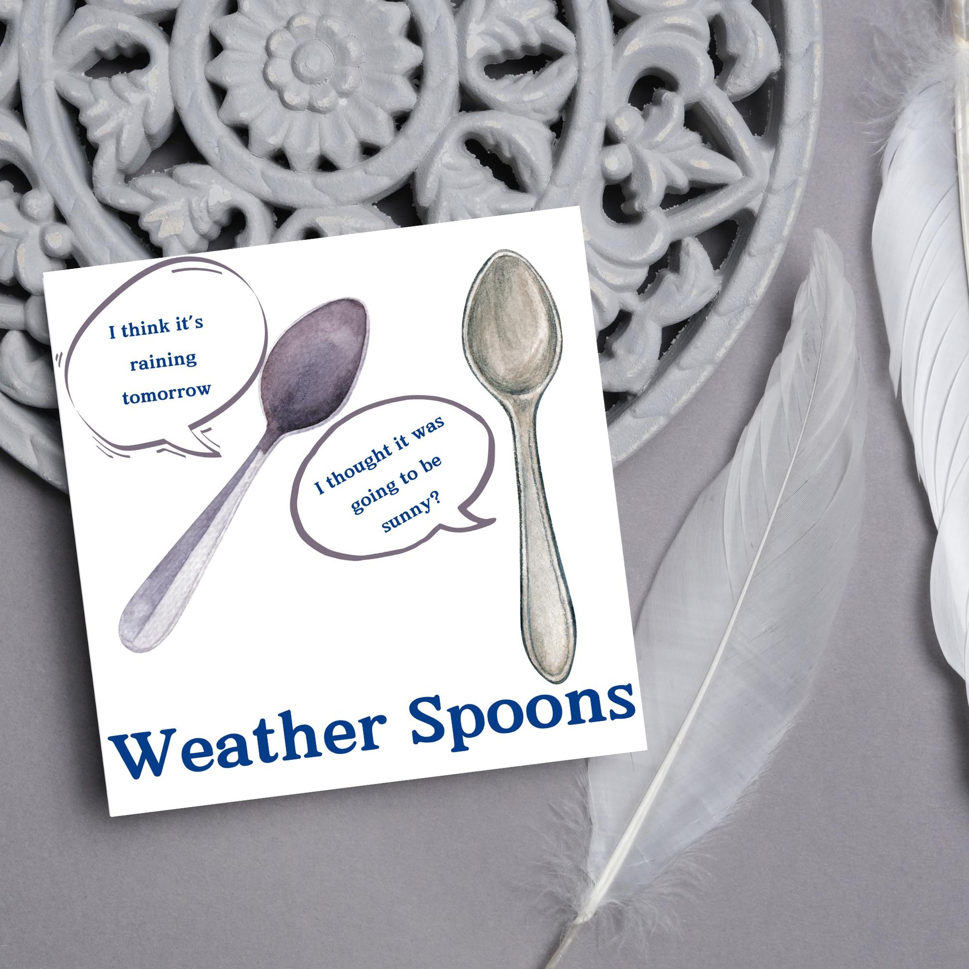 Image of a square greeting card. The card has a white backdrop with an illustration of two spoons with speech bubbles. One says I think it&#39;s raining tomorrow. The other says I thought it was going to be sunny? Underneath the illustration in a blue font is the text Weather Spoons