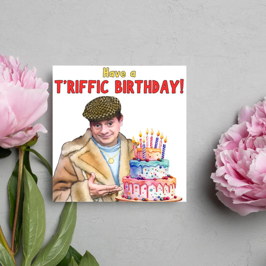 This square shaped card has an illustration of Del Boy from the British Sitcom Only Fools and Horses. The Card has Del Boy standing and pointing at an illustration of a triple tower birthday cake. The Caption reads have a T&#39;riffic Birthday! The Caption is in a mustard yellow and ketchup red