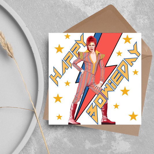 image of a square birthday card. Card has a white background with a vibrant illustration of David bowie with red hair and wearing a striped jumpsuit, behind him is the iconic Bowie lightning bolt in bright red and blue. Diagonal text reads Happy Bowieday in zigzag design bold yellow font. There are yellow stars dotted around the white background.