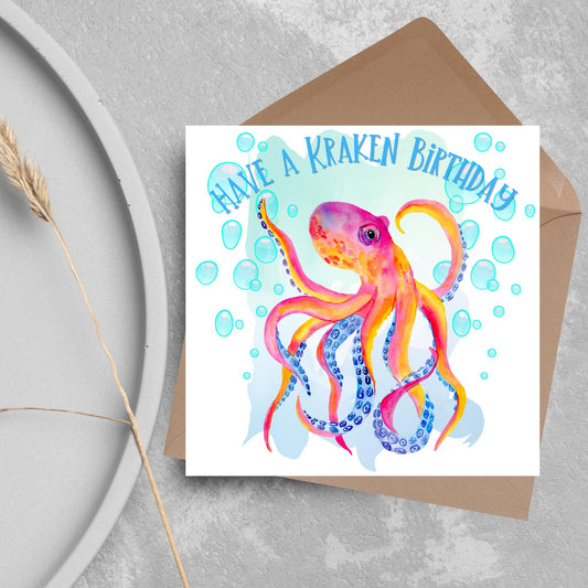 Image of a square birthday card. The background of the card is white and in the centre is a large watercolour image of an octopus in red, orange and blue tones against a light blue bubbly backdrop. Above the Octopus in blue capitalised text, have a kraken birthday is written in a handwritten style font