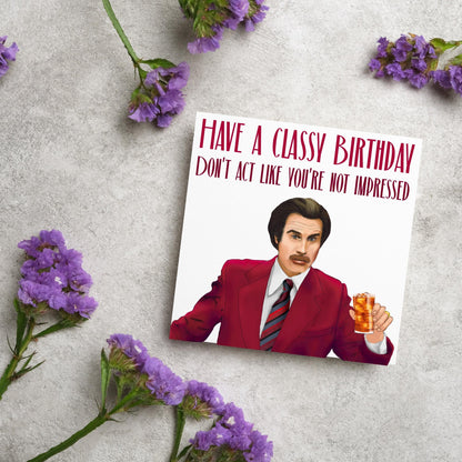 image of a square birthday card, the card has a white background with a hand drawn illustration of Ron Burgandy from the Movie Anchorman  in a dark red suit holding a glass of whiskey. Above him is dark red capitalised text that reads &#39;Have a classy birthday, Don&#39;t act like you&#39;re not impressed.&#39;