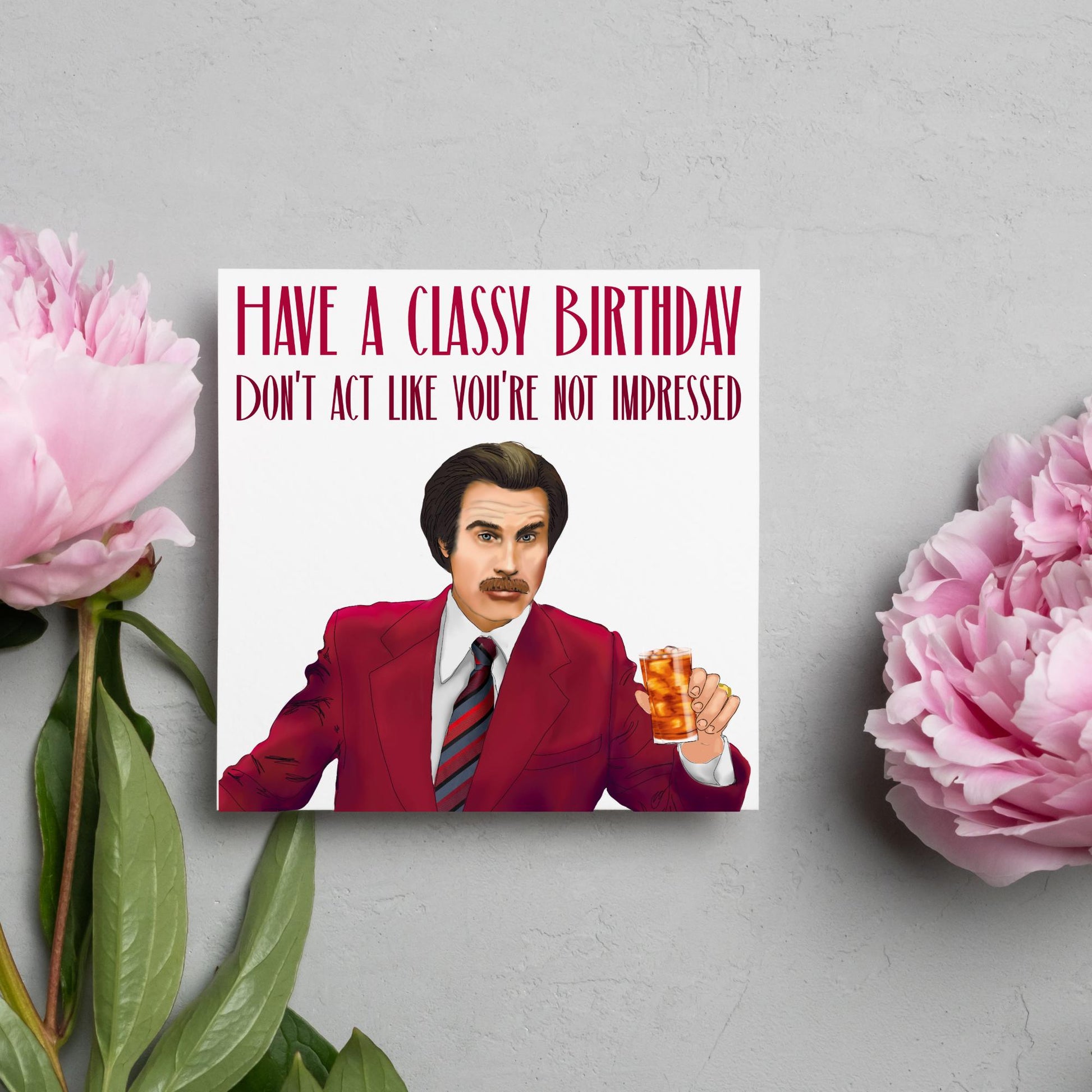 image of a square birthday card, the card has a white background with a hand drawn illustration of Ron Burgandy from the Movie Anchorman  in a dark red suit holding a glass of whiskey. Above him is dark red capitalised text that reads &#39;Have a classy birthday, Don&#39;t act like you&#39;re not impressed.&#39;