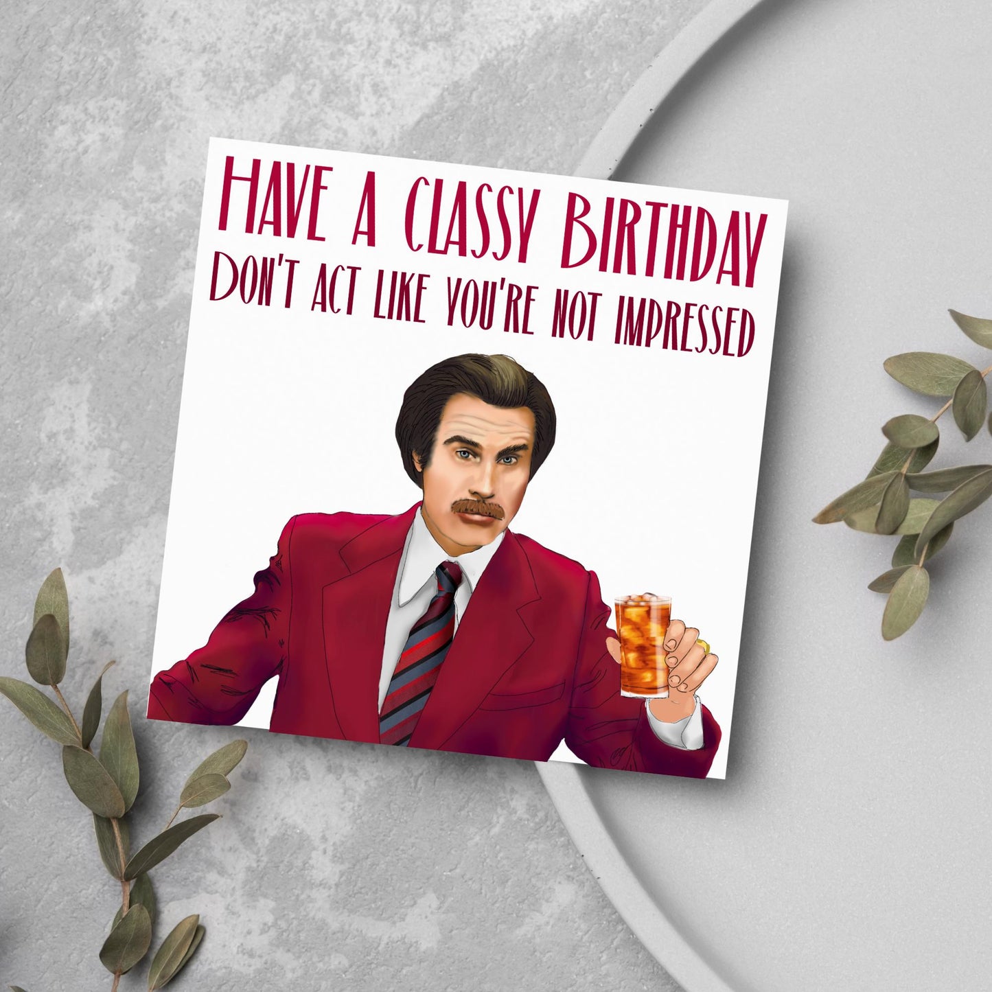image of a square birthday card, the card has a white background with a hand drawn illustration of Ron Burgandy from the Movie Anchorman  in a dark red suit holding a glass of whiskey. Above him is dark red capitalised text that reads &#39;Have a classy birthday, Don&#39;t act like you&#39;re not impressed.&#39;