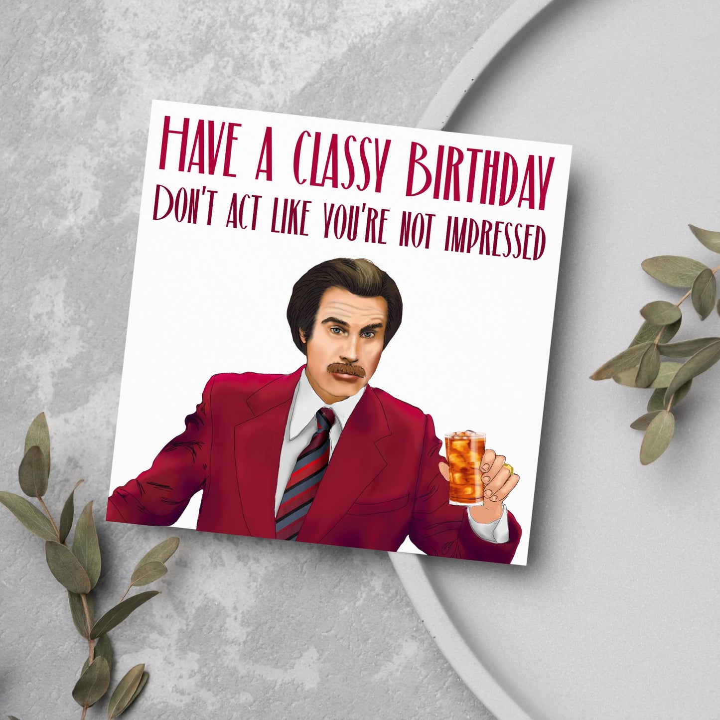 image of a square birthday card, the card has a white background with a hand drawn illustration of Ron Burgandy from the Movie Anchorman  in a dark red suit holding a glass of whiskey. Above him is dark red capitalised text that reads &#39;Have a classy birthday, Don&#39;t act like you&#39;re not impressed.&#39;