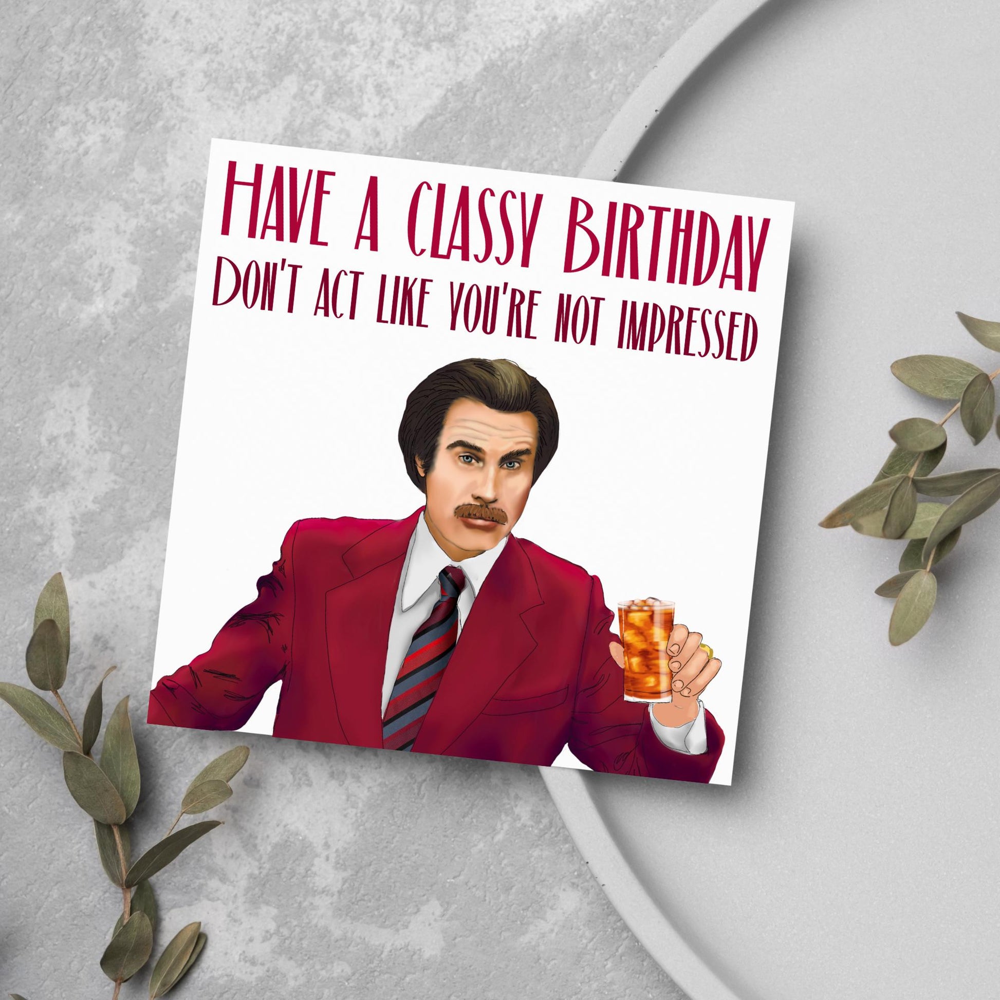 image of a square birthday card, the card has a white background with a hand drawn illustration of Ron Burgandy from the Movie Anchorman  in a dark red suit holding a glass of whiskey. Above him is dark red capitalised text that reads &#39;Have a classy birthday, Don&#39;t act like you&#39;re not impressed.&#39;