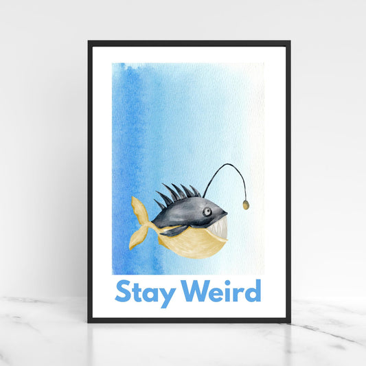 A framed A4 art print featuring a whimsical watercolor illustration of an anglerfish with a yellow tail and light lure, set against a blue gradient background. Below the fish, bold blue text reads &quot;Stay Weird.&quot;