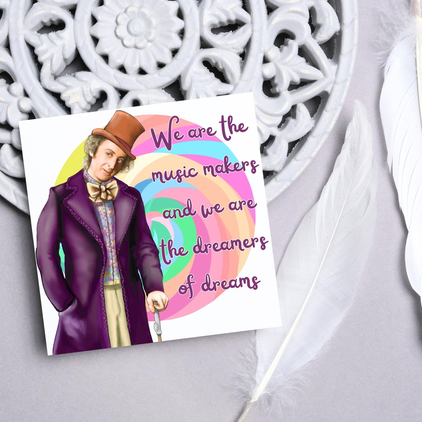 Image of a square greeting card. The background of the card is white and there is a hand drawn image of Gene Wilder as Willy Wonka wearing his purple coat, top hat and bow tie. Behind him is a colourful swirly lollypop and purple text reads &#39;we are the music makers and we are the dreamers of dreams&#39; in a curly font.