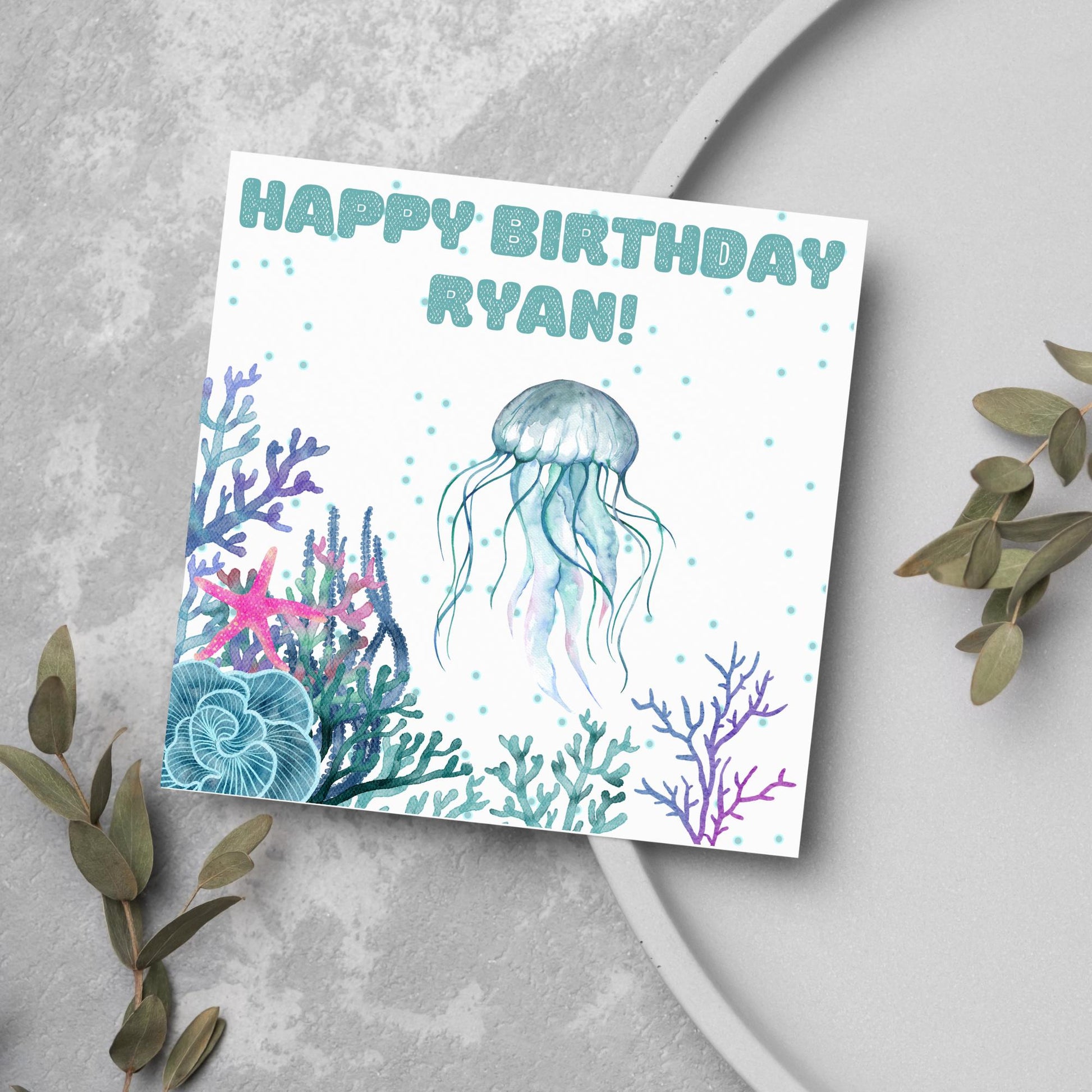 Image of a square Birthday card. Card has a white background and a watercolour style jellyfish amongst coral and seaweed. All the colours are blues, greens, pinks and purples. At the top of the card is bold blue text that says happy birthday ryan which can be personalised to any name.