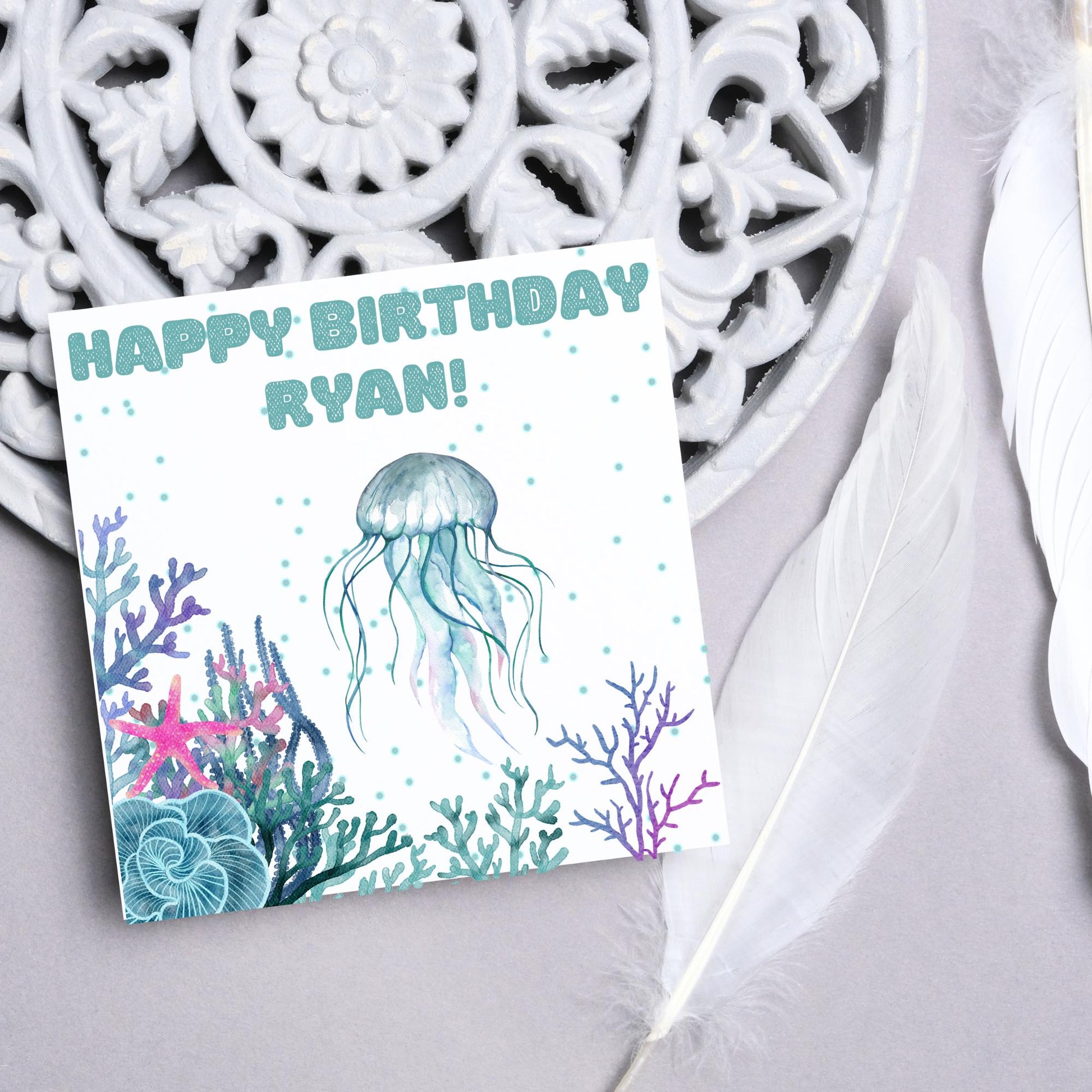 Image of a square Birthday card. Card has a white background and a watercolour style jellyfish amongst coral and seaweed. All the colours are blues, greens, pinks and purples. At the top of the card is bold blue text that says happy birthday ryan which can be personalised to any name.