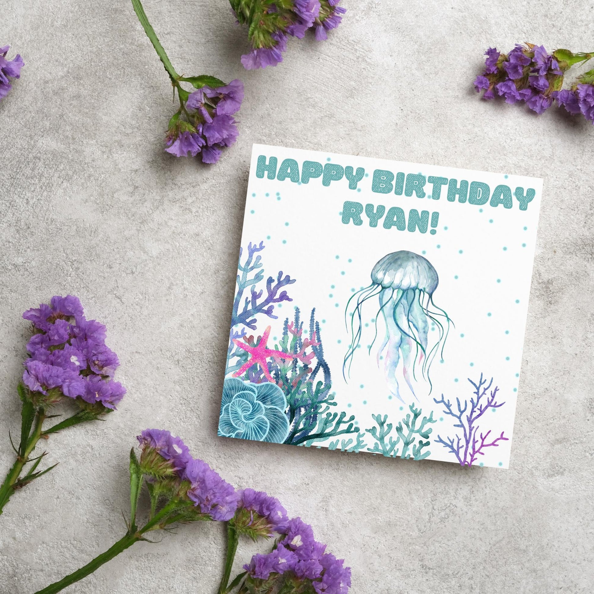 Image of a square Birthday card. Card has a white background and a watercolour style jellyfish amongst coral and seaweed. All the colours are blues, greens, pinks and purples. At the top of the card is bold blue text that says happy birthday ryan which can be personalised to any name.