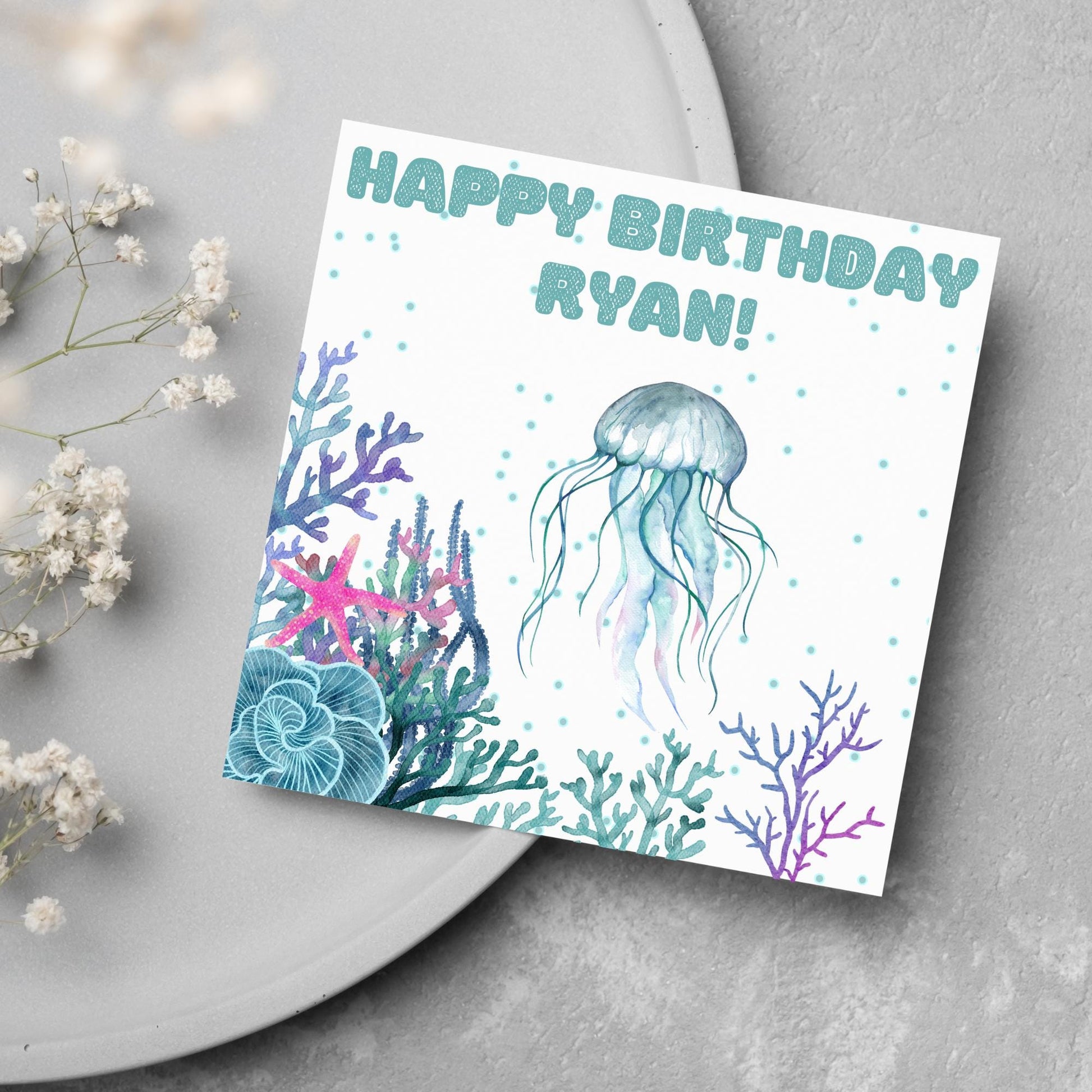Image of a square Birthday card. Card has a white background and a watercolour style jellyfish amongst coral and seaweed. All the colours are blues, greens, pinks and purples. At the top of the card is bold blue text that says happy birthday ryan which can be personalised to any name.