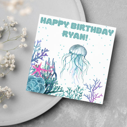 Image of a square Birthday card. Card has a white background and a watercolour style jellyfish amongst coral and seaweed. All the colours are blues, greens, pinks and purples. At the top of the card is bold blue text that says happy birthday ryan which can be personalised to any name.
