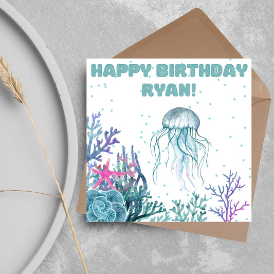 Image of a square Birthday card. Card has a white background and a watercolour style jellyfish amongst coral and seaweed. All the colours are blues, greens, pinks and purples. At the top of the card is bold blue text that says happy birthday ryan which can be personalised to any name.