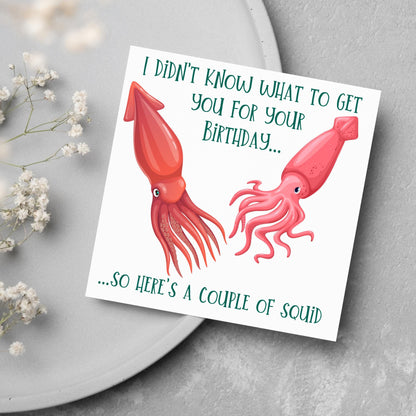Image of a square birthday card. The background of the card is white and there is an illustration of 2 red squid in the centre. Writing surrounds the picture with &#39;I didn&#39;t know what to get you for your birthday....so here&#39;s a couple of squid&#39; in a green capitalised handwritten font.