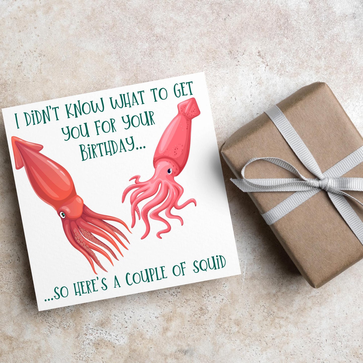 Image of a square birthday card. The background of the card is white and there is an illustration of 2 red squid in the centre. Writing surrounds the picture with &#39;I didn&#39;t know what to get you for your birthday....so here&#39;s a couple of squid&#39; in a green capitalised handwritten font.