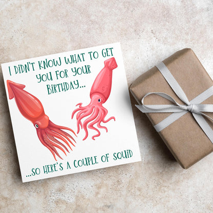 Image of a square birthday card. The background of the card is white and there is an illustration of 2 red squid in the centre. Writing surrounds the picture with &#39;I didn&#39;t know what to get you for your birthday....so here&#39;s a couple of squid&#39; in a green capitalised handwritten font.