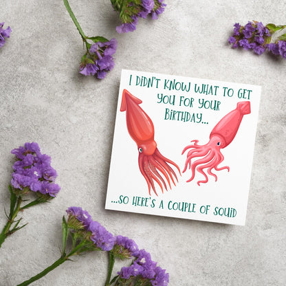 Image of a square birthday card. The background of the card is white and there is an illustration of 2 red squid in the centre. Writing surrounds the picture with &#39;I didn&#39;t know what to get you for your birthday....so here&#39;s a couple of squid&#39; in a green capitalised handwritten font.