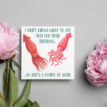 Image of a square birthday card. The background of the card is white and there is an illustration of 2 red squid in the centre. Writing surrounds the picture with &#39;I didn&#39;t know what to get you for your birthday....so here&#39;s a couple of squid&#39; in a green capitalised handwritten font.