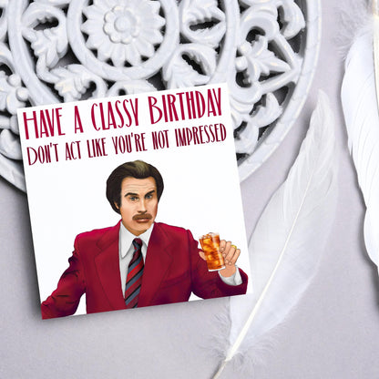 image of a square birthday card, the card has a white background with a hand drawn illustration of Ron Burgandy from the Movie Anchorman  in a dark red suit holding a glass of whiskey. Above him is dark red capitalised text that reads &#39;Have a classy birthday, Don&#39;t act like you&#39;re not impressed.&#39;