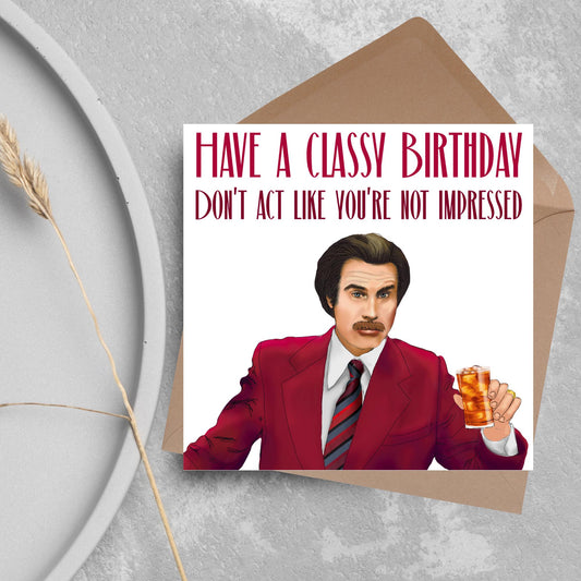 image of a square birthday card, the card has a white background with a hand drawn illustration of Ron Burgandy from the Movie Anchorman  in a dark red suit holding a glass of whiskey. Above him is dark red capitalised text that reads &#39;Have a classy birthday, Don&#39;t act like you&#39;re not impressed.&#39;