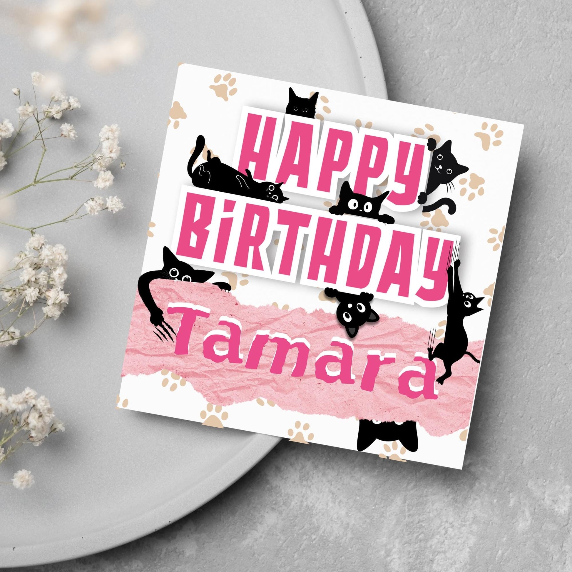 A square birthday card with a white background featuring playful black cartoon cats surrounding and interacting with bold pink text that says &quot;HAPPY BiRTHDAY.&quot; The name &quot;Tamara&quot; is written in a casual, pink handwritten font across a torn paper-style pink banner. Light brown paw prints are scattered in the background.