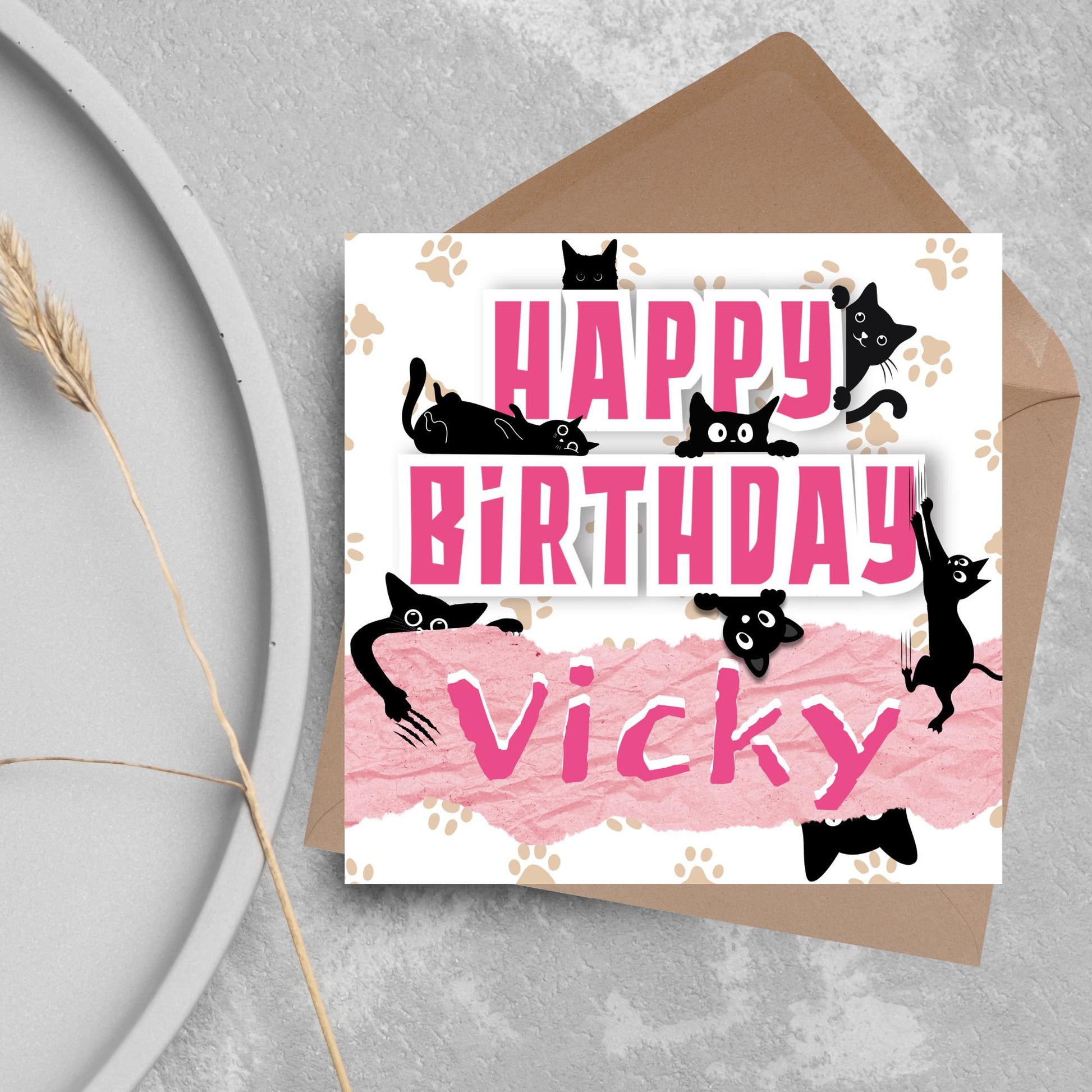 A square birthday card with a white background featuring playful black cartoon cats surrounding and interacting with bold pink text that says &quot;HAPPY BiRTHDAY.&quot; The name &quot;Vicky&quot; is written in a casual, pink handwritten font across a torn paper-style pink banner. Light brown paw prints are scattered in the background. The card is displayed on a gray surface with a brown envelope behind it.