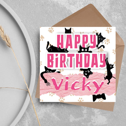 A square birthday card with a white background featuring playful black cartoon cats surrounding and interacting with bold pink text that says &quot;HAPPY BiRTHDAY.&quot; The name &quot;Vicky&quot; is written in a casual, pink handwritten font across a torn paper-style pink banner. Light brown paw prints are scattered in the background. The card is displayed on a gray surface with a brown envelope behind it.
