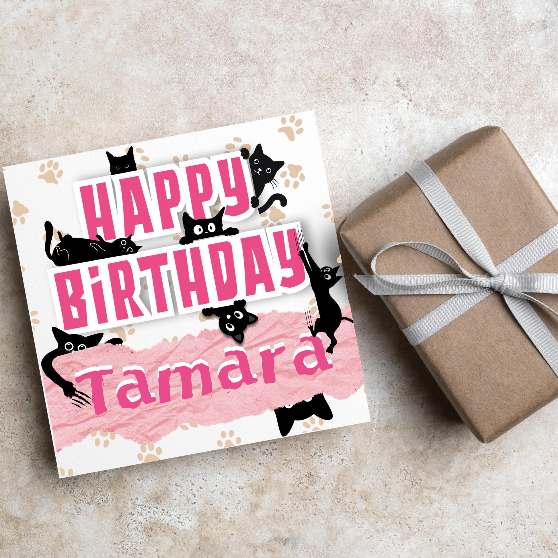 A square birthday card with a white background featuring playful black cartoon cats surrounding and interacting with bold pink text that says &quot;HAPPY BiRTHDAY.&quot; The name &quot;Tamara&quot; is written in a casual, pink handwritten font across a torn paper-style pink banner. Light brown paw prints are scattered in the background.