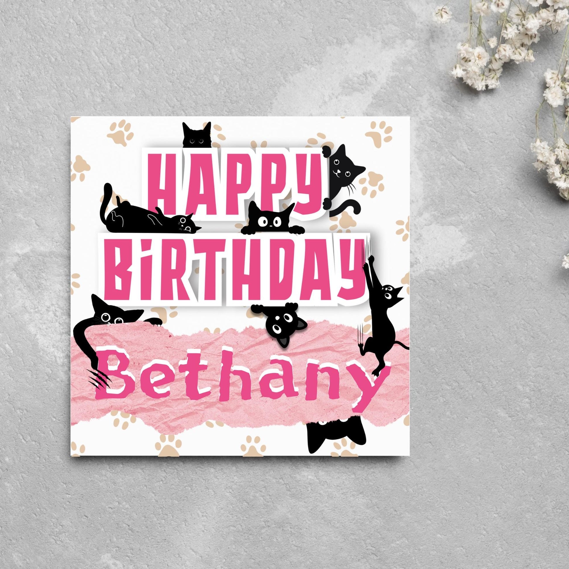 A square birthday card with a white background featuring playful black cartoon cats surrounding and interacting with bold pink text that says &quot;HAPPY BiRTHDAY.&quot; The name &quot;Bethany&quot; is written in a casual, pink handwritten font across a torn paper-style pink banner. Light brown paw prints are scattered in the background.