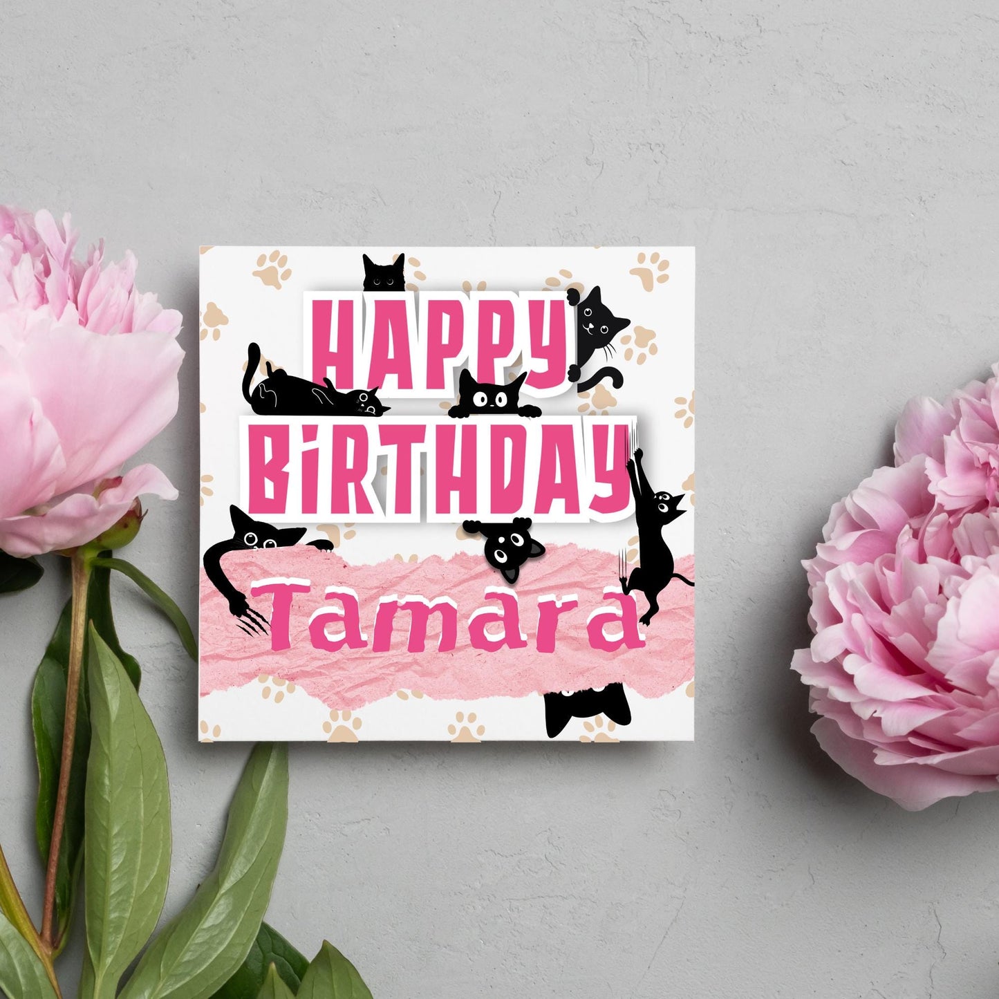 A square birthday card with a white background featuring playful black cartoon cats surrounding and interacting with bold pink text that says &quot;HAPPY BiRTHDAY.&quot; The name &quot;Tamara&quot; is written in a casual, pink handwritten font across a torn paper-style pink banner. Light brown paw prints are scattered in the background.