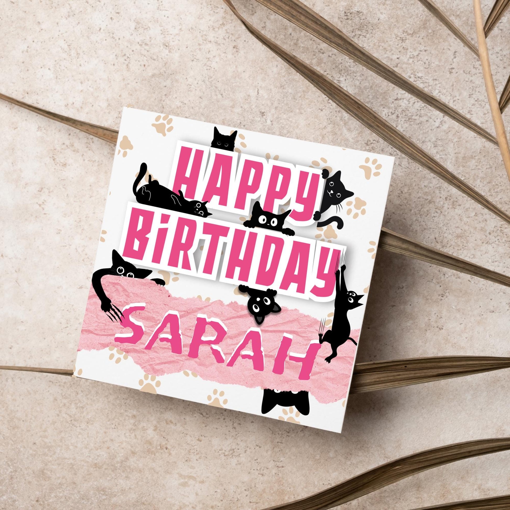 A square birthday card with a white background featuring playful black cartoon cats surrounding and interacting with bold pink text that says &quot;HAPPY BiRTHDAY.&quot; The name &quot;Sarah&quot; is written in a casual, pink handwritten font across a torn paper-style pink banner. Light brown paw prints are scattered in the background.