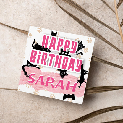A square birthday card with a white background featuring playful black cartoon cats surrounding and interacting with bold pink text that says &quot;HAPPY BiRTHDAY.&quot; The name &quot;Sarah&quot; is written in a casual, pink handwritten font across a torn paper-style pink banner. Light brown paw prints are scattered in the background.