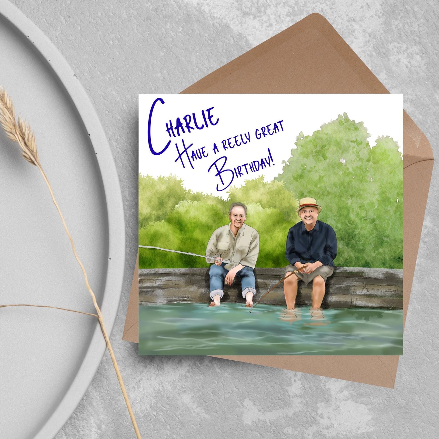 square greetings card with a hand drawn image of bob Mortimer and paul Whitehouse sitting on the edge of a dock fishing with their bare feet in the water. behind them are watercolour trees and bushes and blue text that reads &#39;charlie, have a freely great birthday&#39; in a handwritten style font