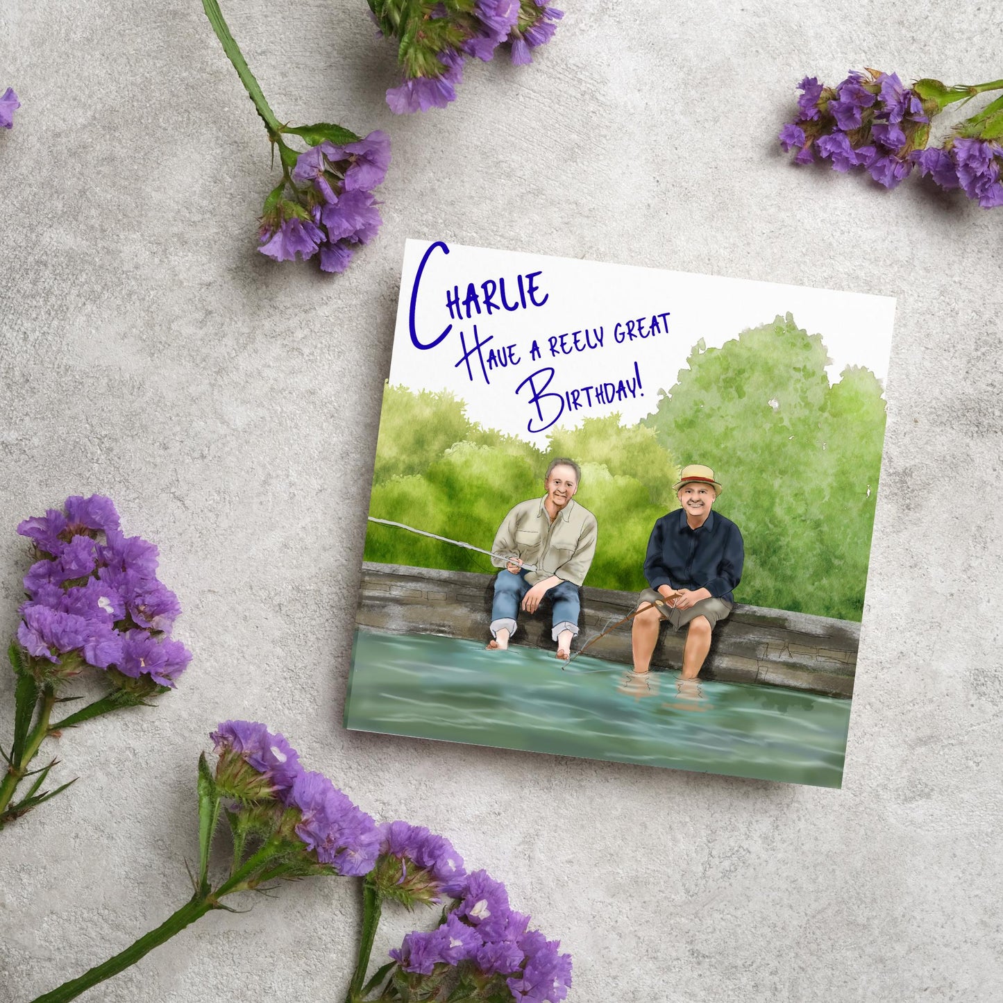 square greetings card with a hand drawn image of bob Mortimer and paul Whitehouse sitting on the edge of a dock fishing with their bare feet in the water. behind them are watercolour trees and bushes and blue text that reads &#39;charlie, have a freely great birthday&#39; in a handwritten style font