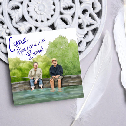 square greetings card with a hand drawn image of bob Mortimer and paul Whitehouse sitting on the edge of a dock fishing with their bare feet in the water. behind them are watercolour trees and bushes and blue text that reads &#39;charlie, have a freely great birthday&#39; in a handwritten style font