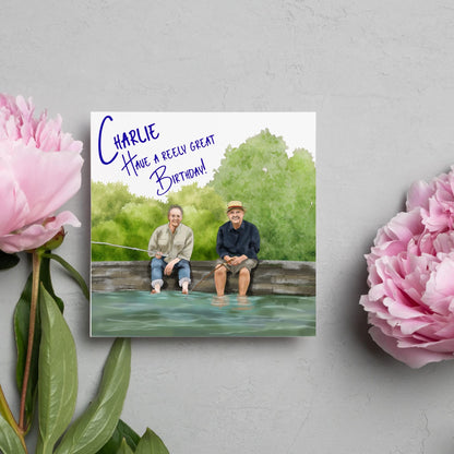 square greetings card with a hand drawn image of bob Mortimer and paul Whitehouse sitting on the edge of a dock fishing with their bare feet in the water. behind them are watercolour trees and bushes and blue text that reads &#39;charlie, have a freely great birthday&#39; in a handwritten style font