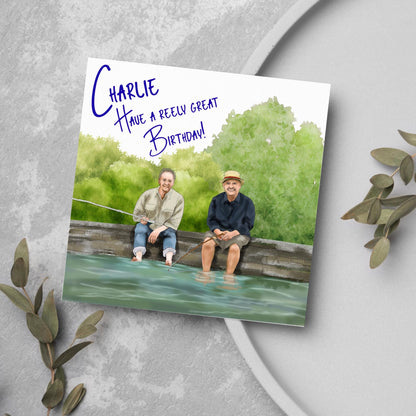 square greetings card with a hand drawn image of bob Mortimer and paul Whitehouse sitting on the edge of a dock fishing with their bare feet in the water. behind them are watercolour trees and bushes and blue text that reads &#39;charlie, have a freely great birthday&#39; in a handwritten style font